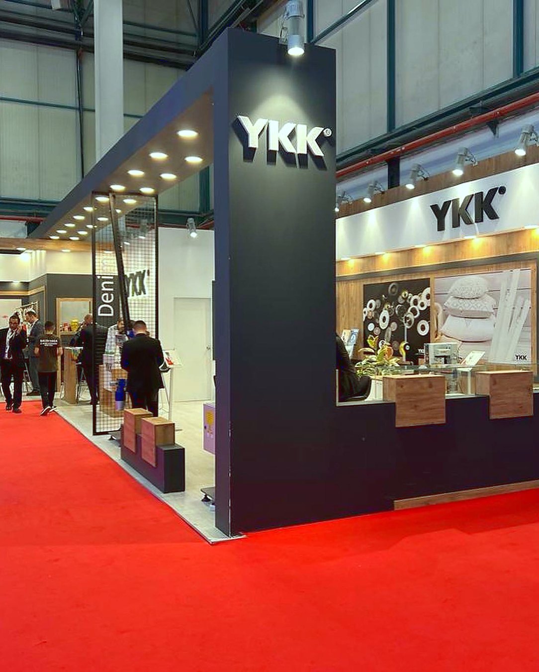 Stand d’exposition YKK au salon IGM 2022 : Une tour aux tons bois, des panneaux produits ordonnés et un espace de réunion compact structurent une gamme étendue